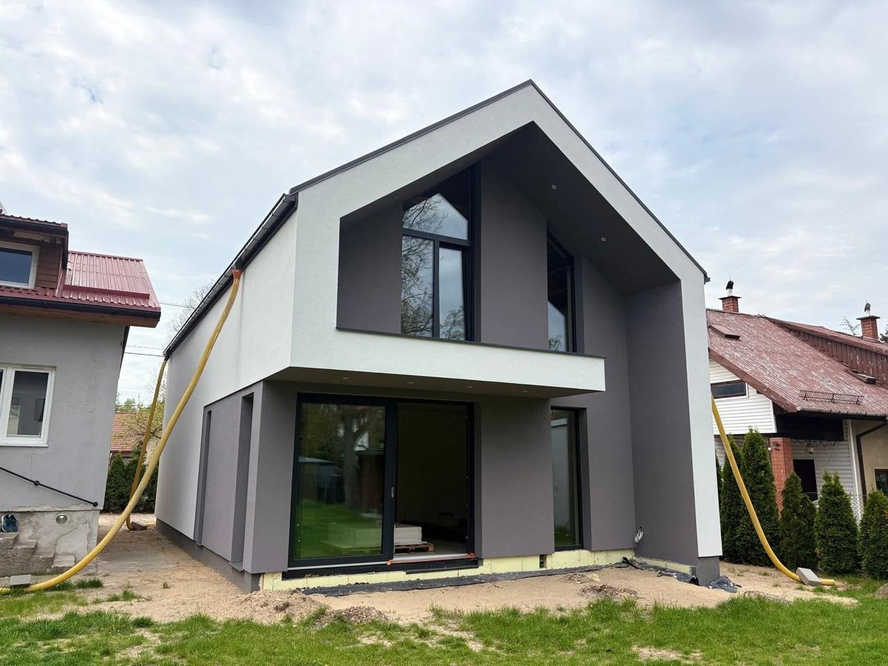 Nowoczesny dom z elewacją w odcieniach szarości i bieli, z widocznymi elementami ocieplenia i żółtymi rurami wentylacyjnymi, na tle pochmurnego nieba.