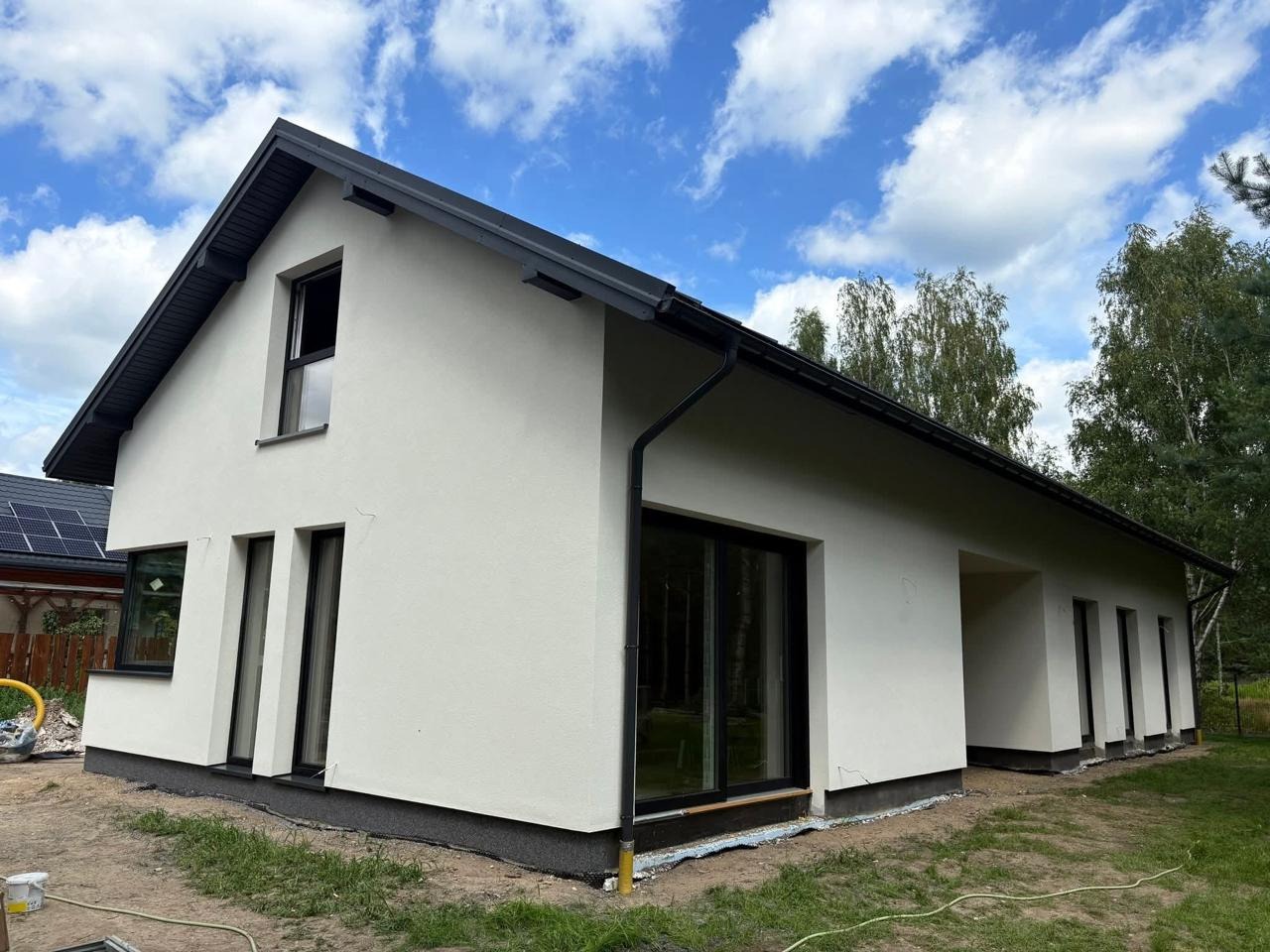 Nowoczesny dom jednorodzinny z białą elewacją i czarnymi oknami, widok z zewnątrz w słoneczny dzień. Minimalistyczny design, czyste linie, otoczenie zieleni.