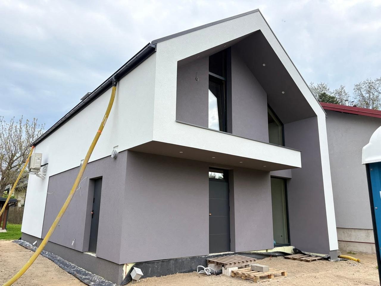 Nowoczesny dom po ociepleniu ścian zewnętrznych, z widoczną żółtą rurą do wdmuchiwania izolacji. Elewacja w odcieniach szarości i bieli, minimalistyczny design.