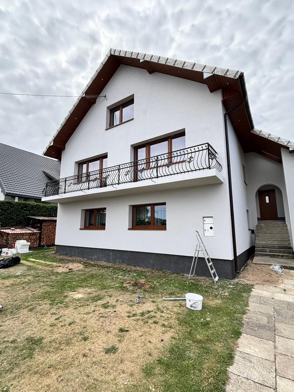 Biała elewacja domu z balkonem i brązowymi oknami po renowacji. Widoczna drabina, wiadro z farbą i fragment ogrodu. Dom z poddaszem użytkowym.
