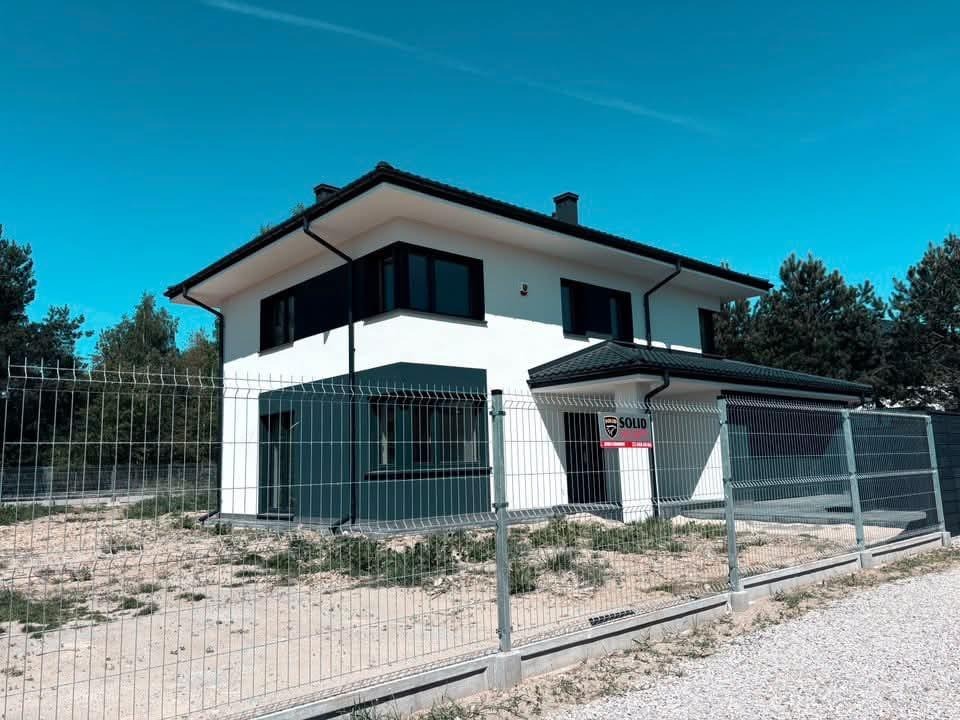 Dwukondygnacyjny dom z białą elewacją i czarnymi akcentami, otoczony metalowym ogrodzeniem panelowym na tle błękitnego nieba i zieleni drzew. Widoczny znak firmy na ogrodzeniu.