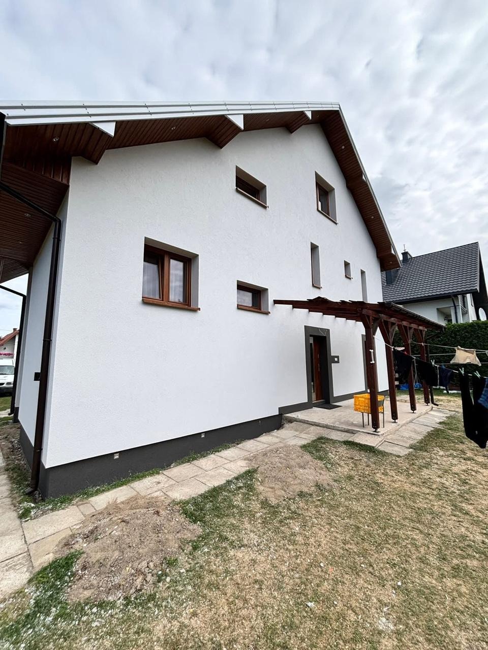 Biała elewacja domu jednorodzinnego z drewnianymi elementami wykończeniowymi, widok z boku na tle pochmurnego nieba i fragmentu ogrodu.