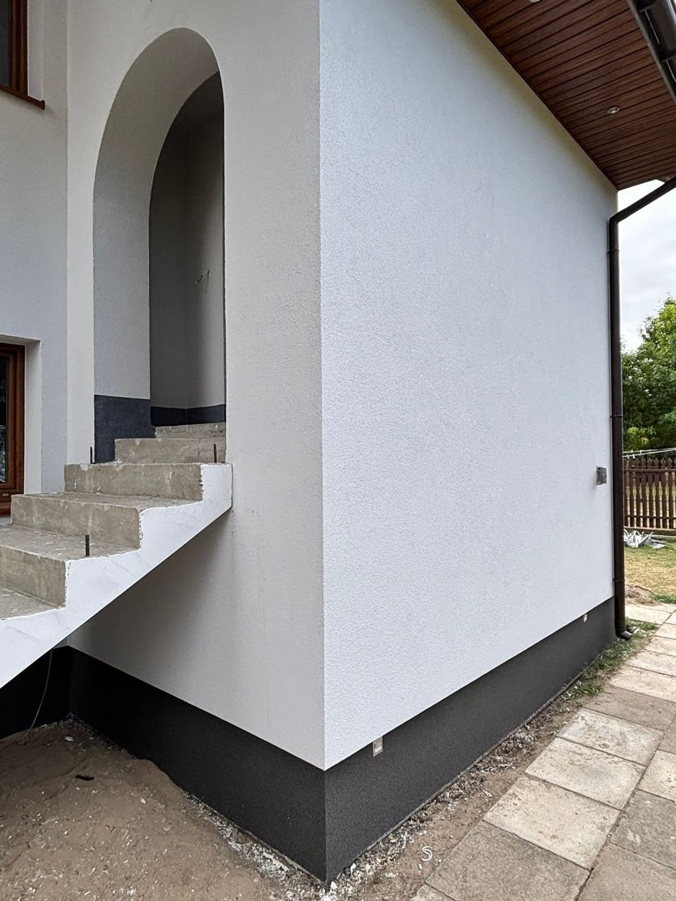 Biała elewacja domu z ciemną podmurówką i łukowym wejściem. Widoczne schody i fragment chodnika. Elewacja o fakturze baranka, świeżo po wykonaniu.
