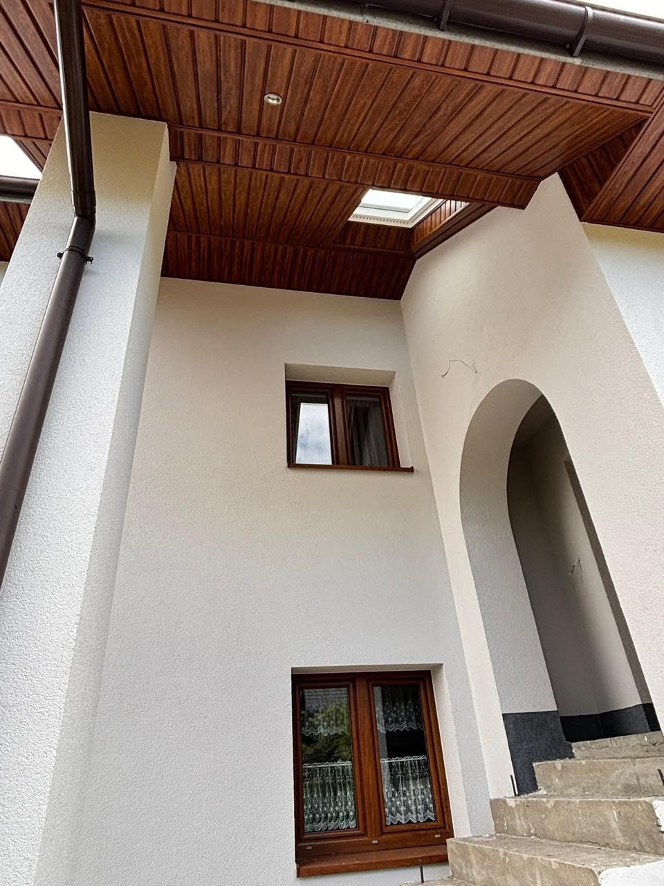 Elewacja domu z oknami i drewnianą podbitką dachową, widok z dołu. Jasna fasada, brązowe okna i rynny. Schody prowadzące do łukowatego wejścia.