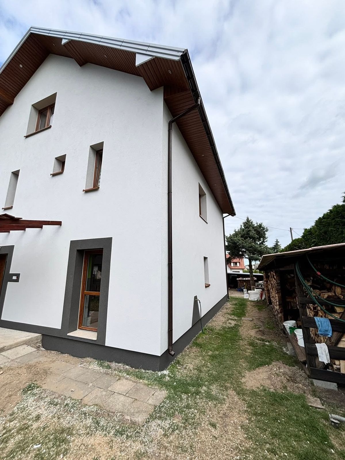Biała elewacja domu jednorodzinnego z brązowymi detalami (okna, rynny, podbitka). Widok z boku, na tle pochmurnego nieba i zieleni. Świeżo pomalowana ściana.