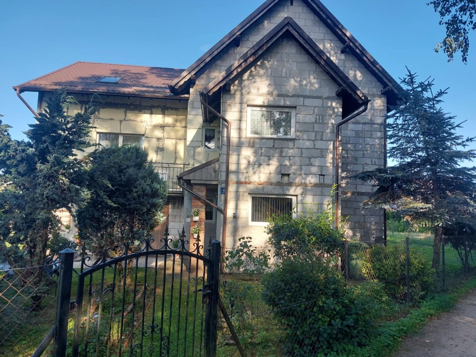 Dwukondygnacyjny dom w trakcie ocieplania: jedna ściana z szarego bloczka, druga z widoczną warstwą izolacyjną. Metalowe ogrodzenie i zieleń na pierwszym planie.