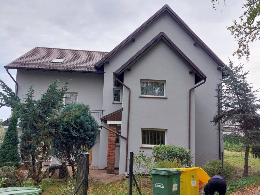Dom jednorodzinny po remoncie elewacji, z nowymi oknami i rynnami w kolorze brązowym. Widoczne kosze na śmieci i fragment osoby w ogrodzie.