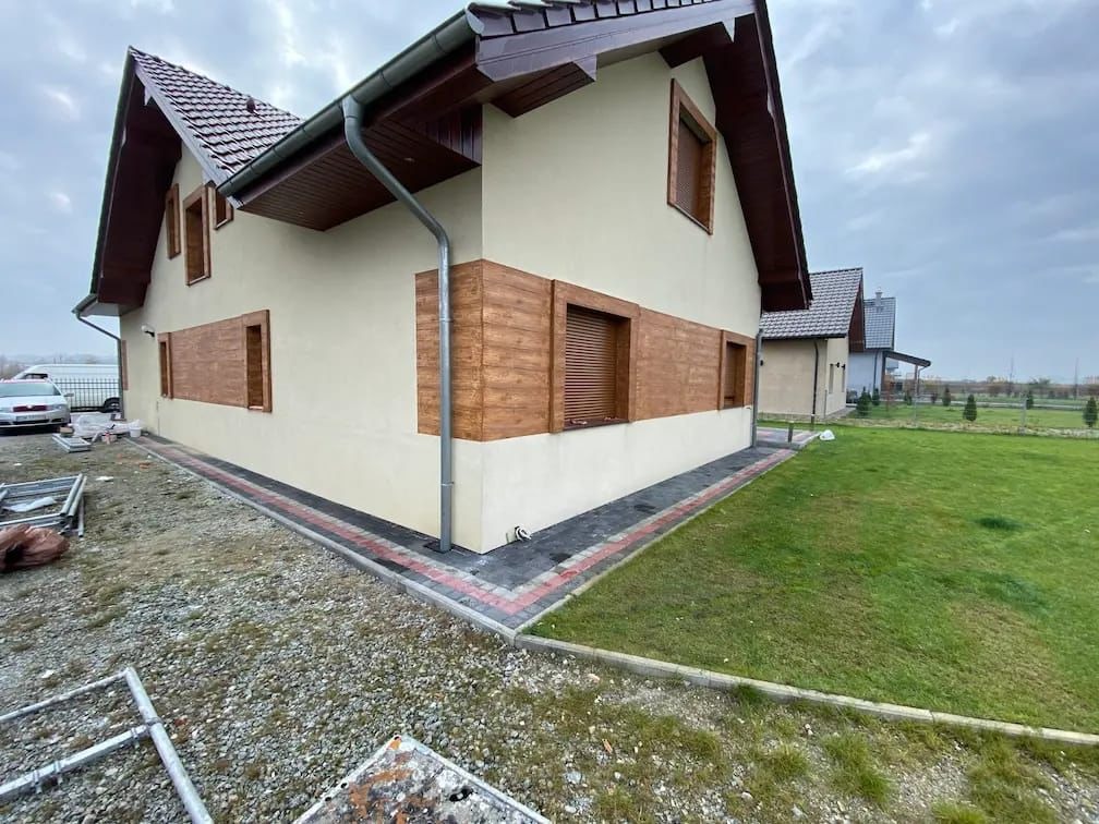 Wykończony dom z beżową elewacją i drewnianymi akcentami. Widoczne okna z roletami, brązowe rynny i fragment ogrodu. Ujęcie z niskiej perspektywy, podkreślające detale wykończenia.