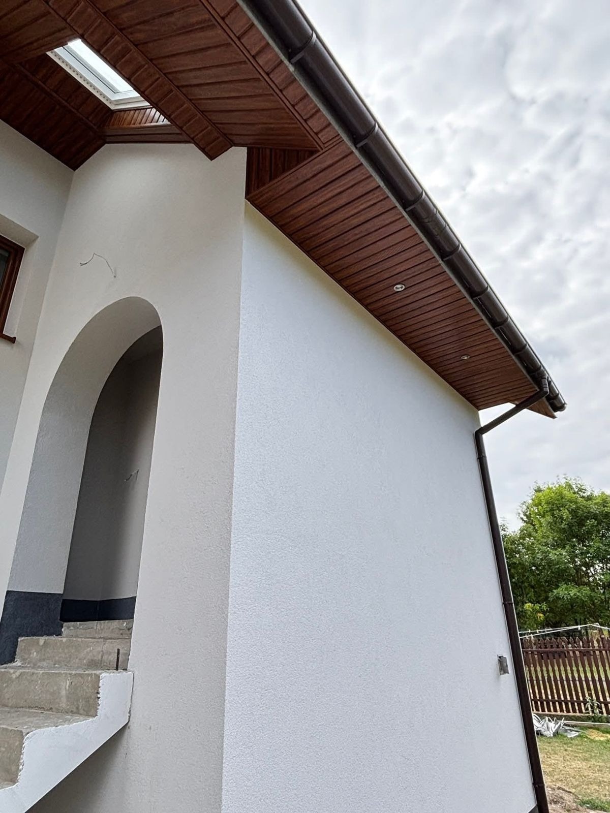Biała elewacja budynku z drewnianą podbitką i brązowymi rynnami, widok z dołu, łukowate wejście, fragment schodów, pochmurne niebo w tle.