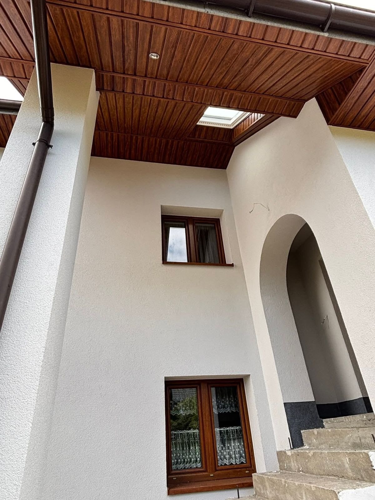 Elewacja domu z oknami w brązowych ramach, widok z dołu na drewnianą podbitkę dachową i fragment rynny. Biała ściana z fakturą.