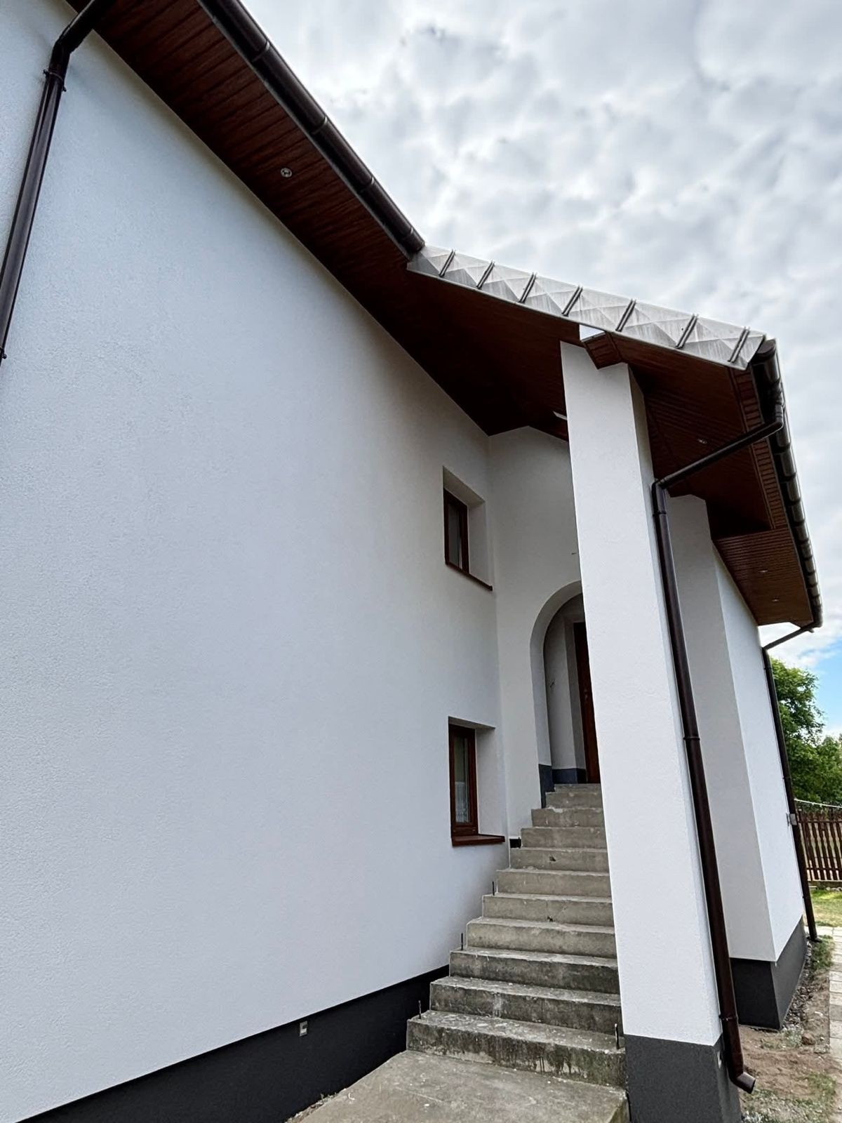 Biała elewacja domu z brązowymi oknami i dachem, widok z dołu, schody wejściowe z łukowym wejściem, rynny w kolorze dachu, fragment ogrodzenia w tle.