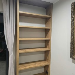 Mistrzowski Wymiar - Wysoka, drewniana szafa wnękowa z półkami, widoczna z boku, z dekoracyjną ramą obrazu w tle. Naturalne oświetlenie podkreśla strukturę drewna.