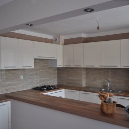 Mistrzowski Wymiar - Jasna kuchnia z drewnianym blatem i beżowymi płytkami. Widoczna płyta gazowa, zlew i dekoracje na blacie. Górne szafki białe, bez uchwytów. Oświetlenie sufitowe.