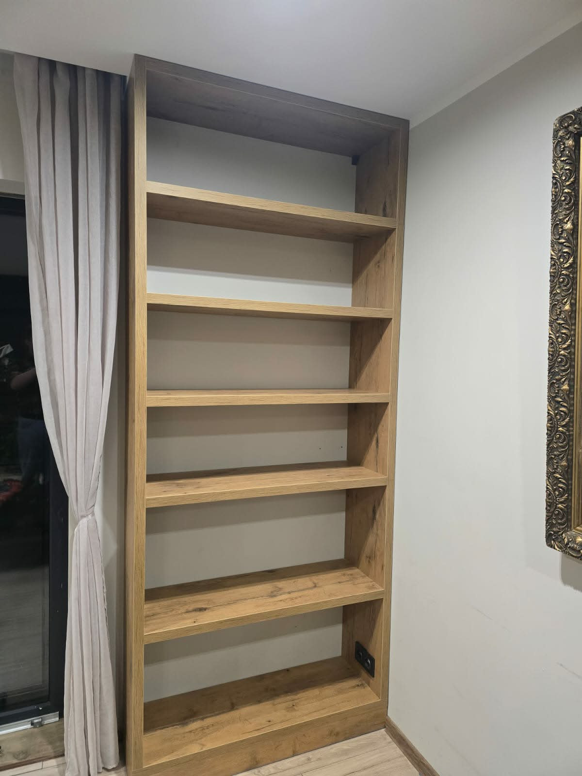 Wysoka, drewniana szafa wnękowa z półkami, widoczna z boku, z dekoracyjną ramą obrazu w tle. Naturalne oświetlenie podkreśla strukturę drewna.