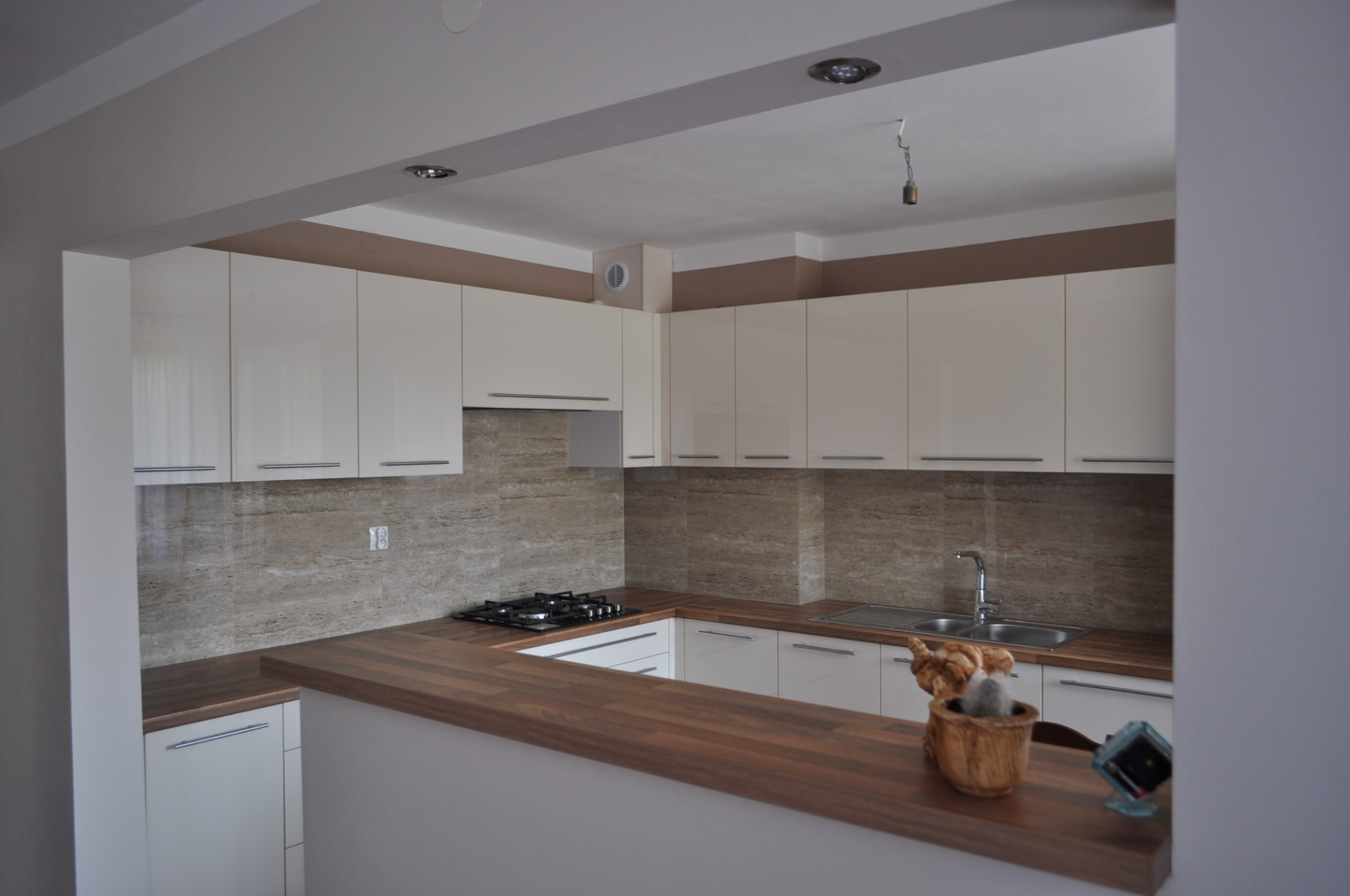 Jasna kuchnia z drewnianym blatem i beżowymi płytkami. Widoczna płyta gazowa, zlew i dekoracje na blacie. Górne szafki białe, bez uchwytów. Oświetlenie sufitowe.