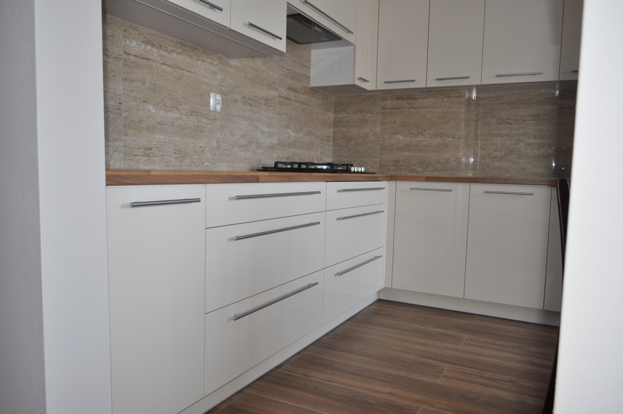 Biała, lakierowana kuchnia z drewnianym blatem i płytkami imitującymi kamień na ścianie. Minimalistyczne uchwyty, płyta gazowa. Widok na narożnik zabudowy.