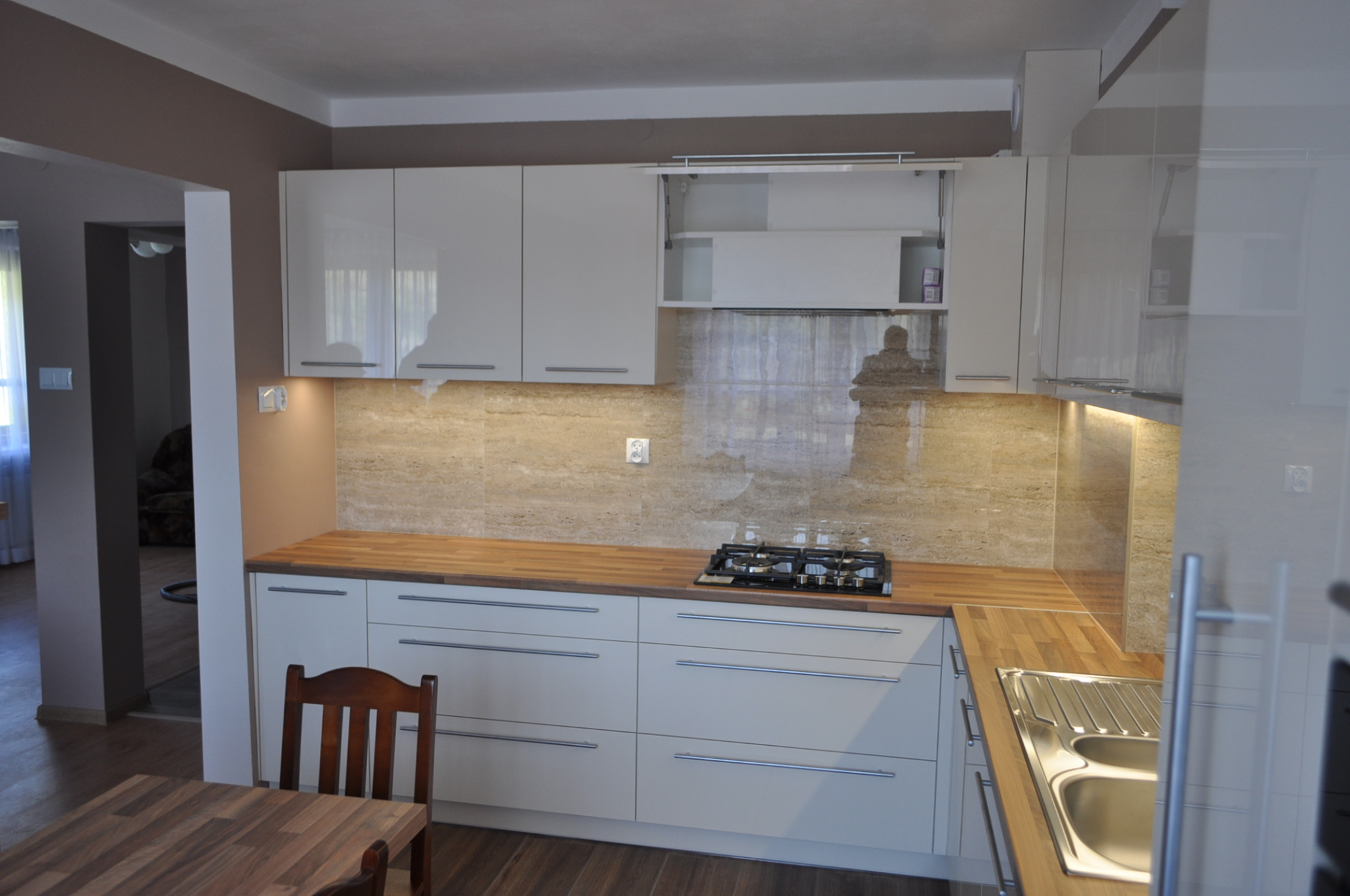 Jasna kuchnia z drewnianym blatem i płytkami imitującymi kamień. Widoczna płyta gazowa i zlew. Krzesło przy stole w tle. Ujęcie z perspektywy.