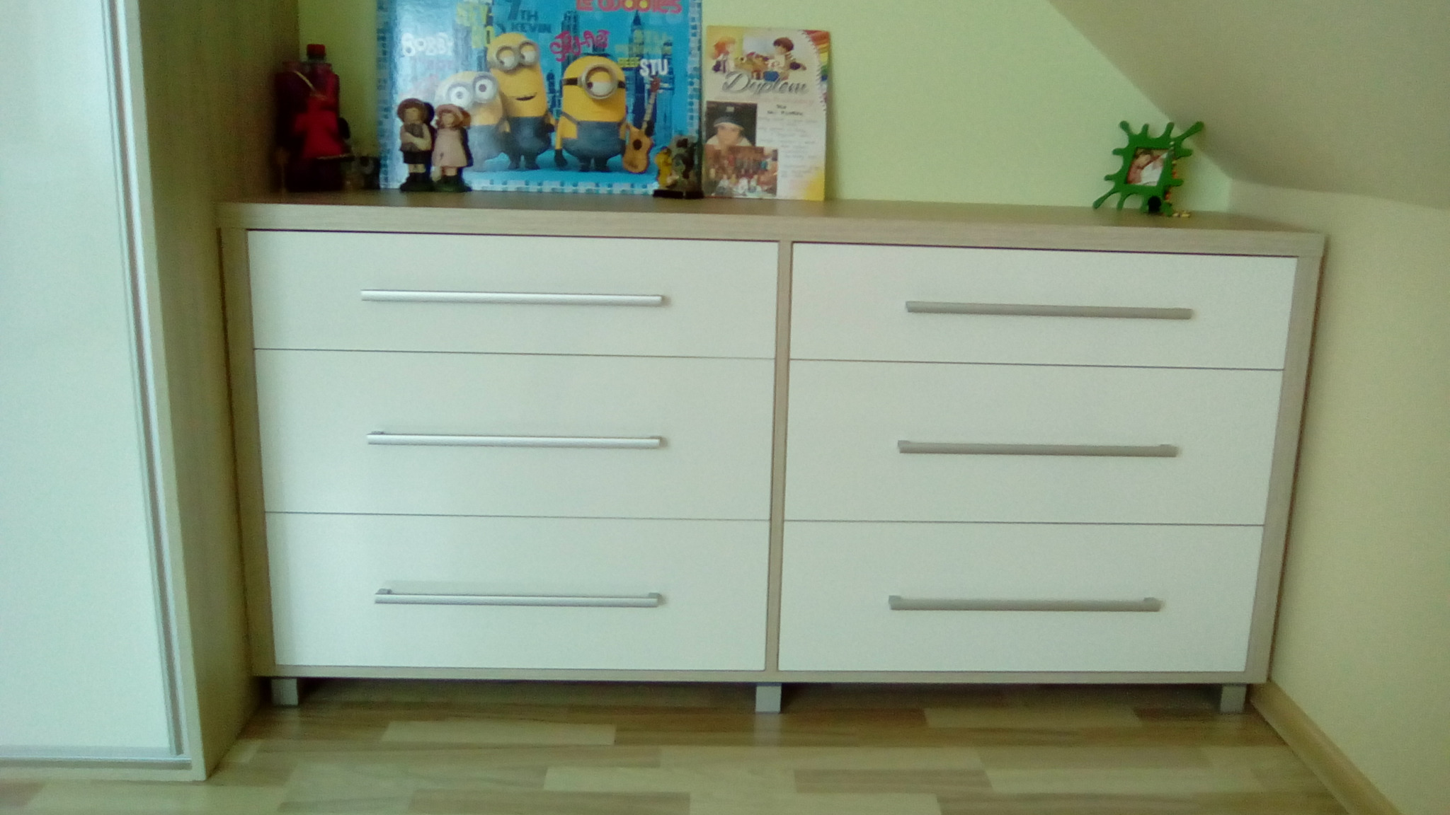 Komoda z sześcioma szufladami w jasnej kolorystyce, z dekoracjami na blacie, ustawiona na drewnianej podłodze, widoczna z boku szafa przesuwna.