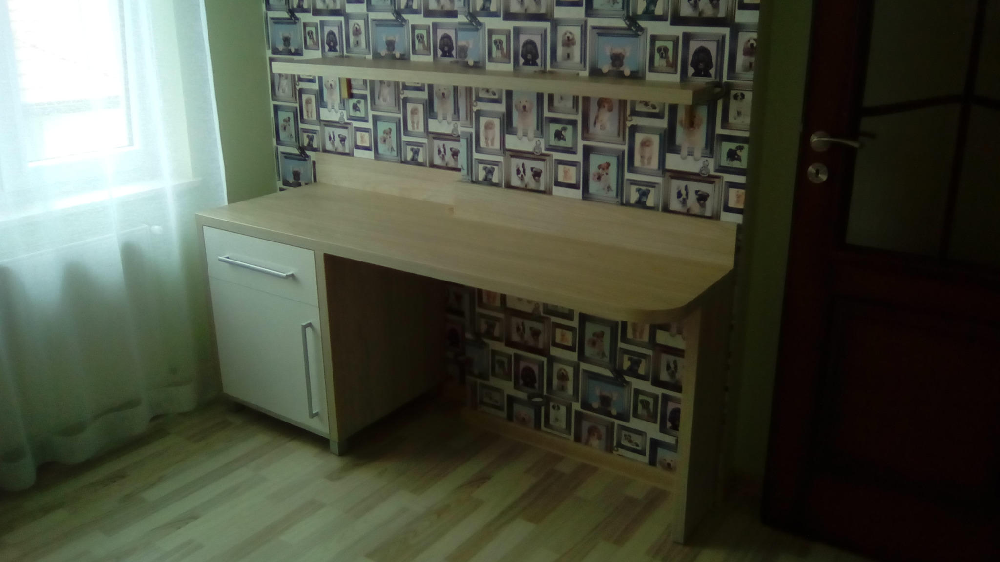 Biurko na wymiar z szafką i półką, jasne drewno, ściana w tapetę z psami w ramkach. Widok z boku, z oknem w tle. Nowoczesny design.