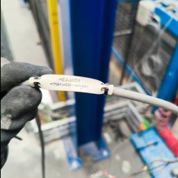 PowerNova-Elektromontaż - Oznakowany przewód w dłoni w rękawicy roboczej na tle konstrukcji. Etykieta z oznaczeniem '=E2-WD1 +77052.UC30/=E2-XG1' na kablu.