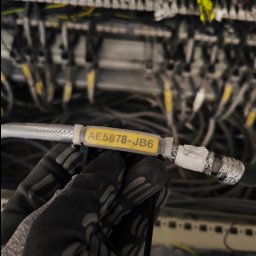 PowerNova-Elektromontaż - Oznakowany przewód koncentryczny w dłoni w rękawicy roboczej na tle szafy telekomunikacyjnej z okablowaniem. Etykieta z kodem 'AE5878-JB6'.