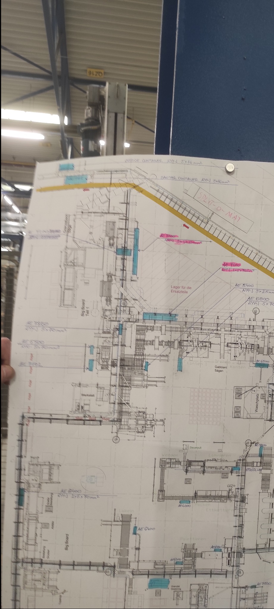 Szkic instalacji elektrycznej z naniesionymi oznaczeniami, widoczny fragment hali przemysłowej w tle. Plan instalacji z detalami technicznymi i oznaczeniami.