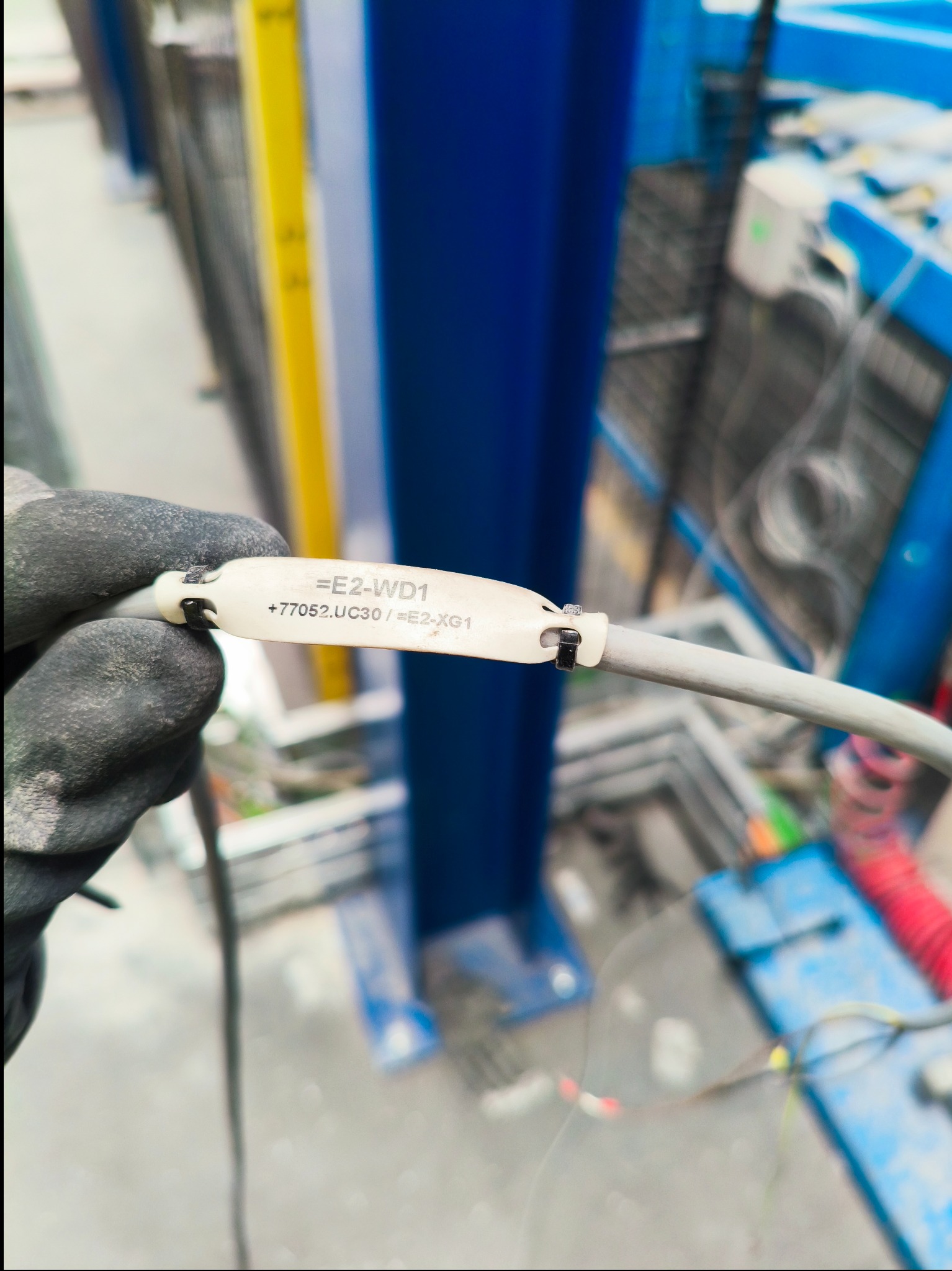 Oznakowany przewód w dłoni w rękawicy roboczej na tle konstrukcji. Etykieta z oznaczeniem '=E2-WD1 +77052.UC30/=E2-XG1' na kablu.
