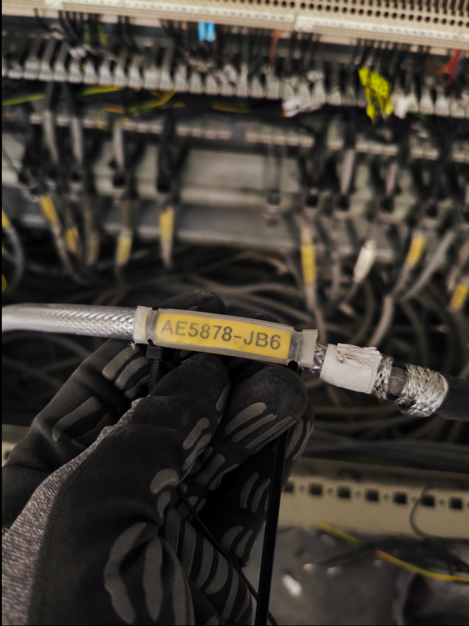 Oznakowany przewód koncentryczny w dłoni w rękawicy roboczej na tle szafy telekomunikacyjnej z okablowaniem. Etykieta z kodem 'AE5878-JB6'.