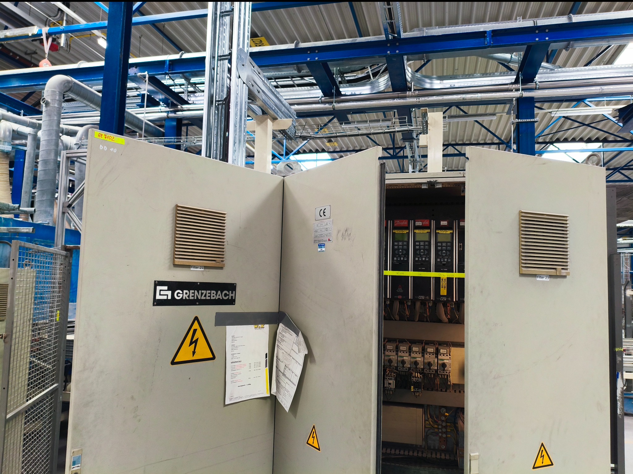 Otwarta szafa sterownicza z widocznymi elementami elektrycznymi, wentylacją i ostrzeżeniami o napięciu. Widoczne kable i aparatura wewnątrz. Metalowa konstrukcja w tle.