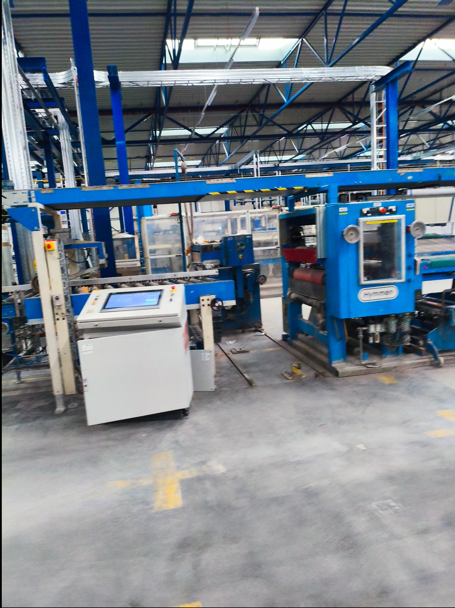 Niebieska linia produkcyjna z panelem sterowania w hali fabrycznej. Widoczne elementy konstrukcyjne i oświetlenie sufitowe. Żółte oznaczenia na podłodze.