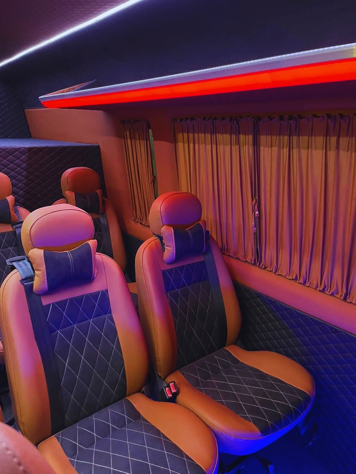 Luksusowe wnętrze busa z brązowo-czarnymi skórzanymi fotelami, pikowaną tapicerką i nastrojowym oświetleniem LED. Zasłony w oknach dodają prywatności.