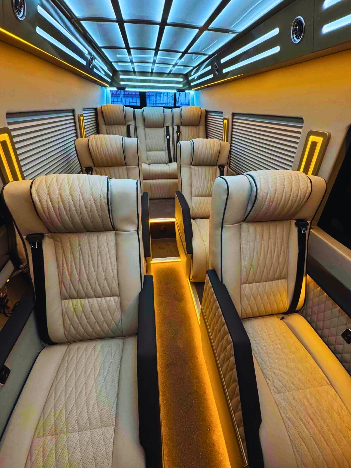 Luksusowe wnętrze busa z beżowymi skórzanymi fotelami, oświetleniem LED na suficie i podłodze, tworzące ekskluzywną atmosferę. Widok na komfortowe siedzenia i detale wykończenia.