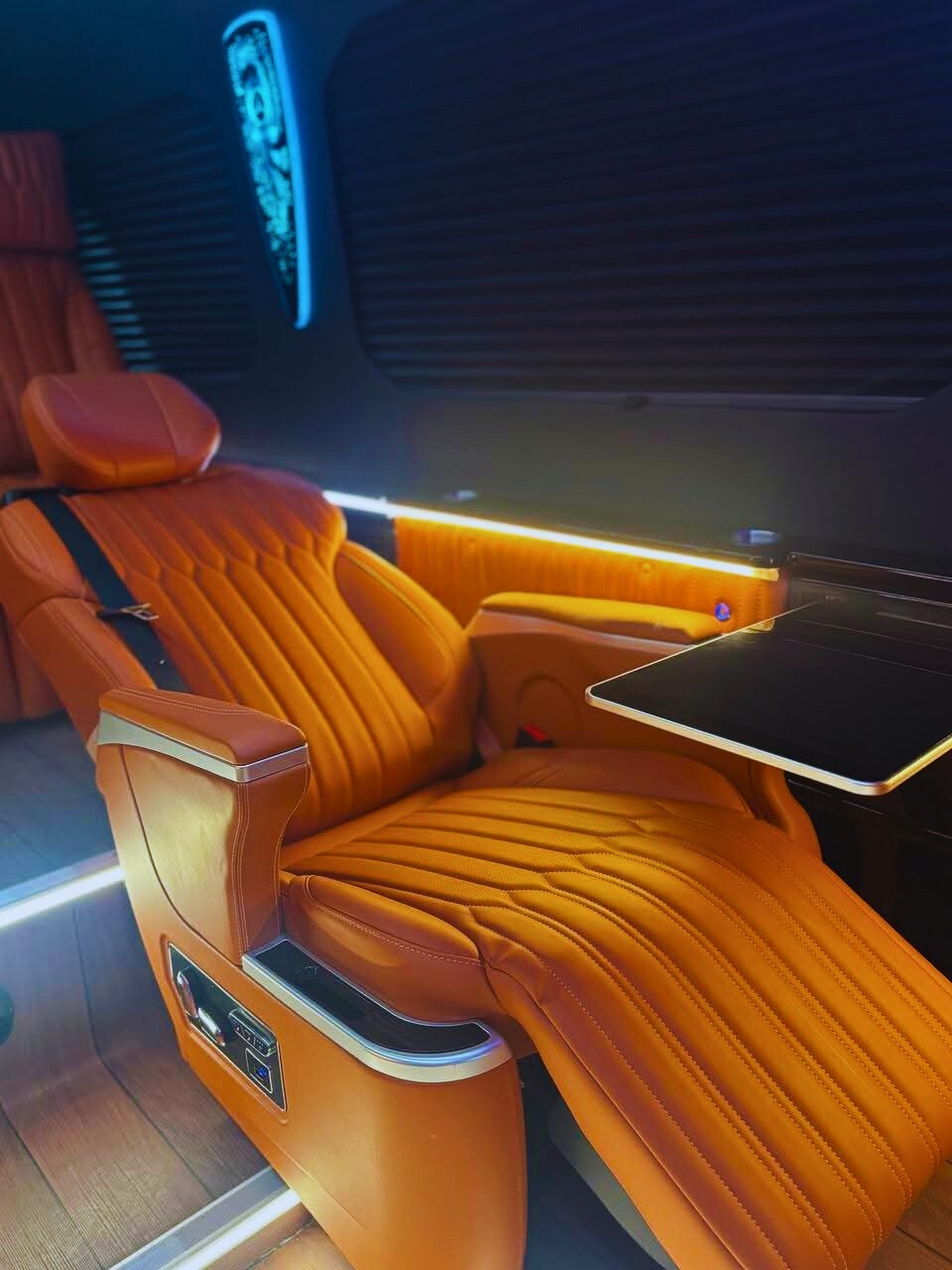 Luksusowy, pomarańczowy fotel skórzany z rozkładanym stolikiem wewnątrz pojazdu. Podświetlenie LED, rolety w oknach. Detal wnętrza busa VIP.