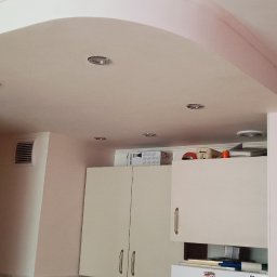 Patryk Kot - Wykończony sufit podwieszany z zaokrąglonym łukiem i wbudowanymi punktami świetlnymi w jasnym wnętrzu kuchni. Widoczne szafki kuchenne i fragment drzwi.