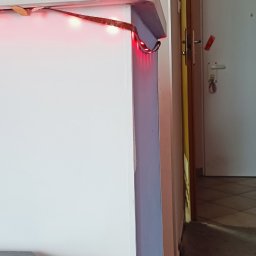 Patryk Kot - Biała ściana z czerwoną listwą LED w rogu pokoju. Widoczny fragment przedpokoju z szafką na buty i ubrania. Ujęcie z bliska, oświetlenie mieszane.