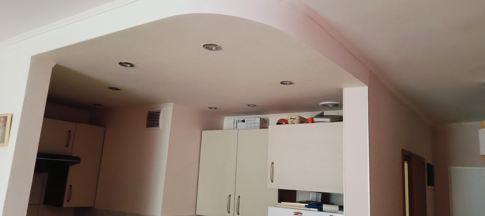 Wykończony sufit podwieszany z zaokrąglonym łukiem i wbudowanymi punktami świetlnymi w jasnym wnętrzu kuchni. Widoczne szafki kuchenne i fragment drzwi.
