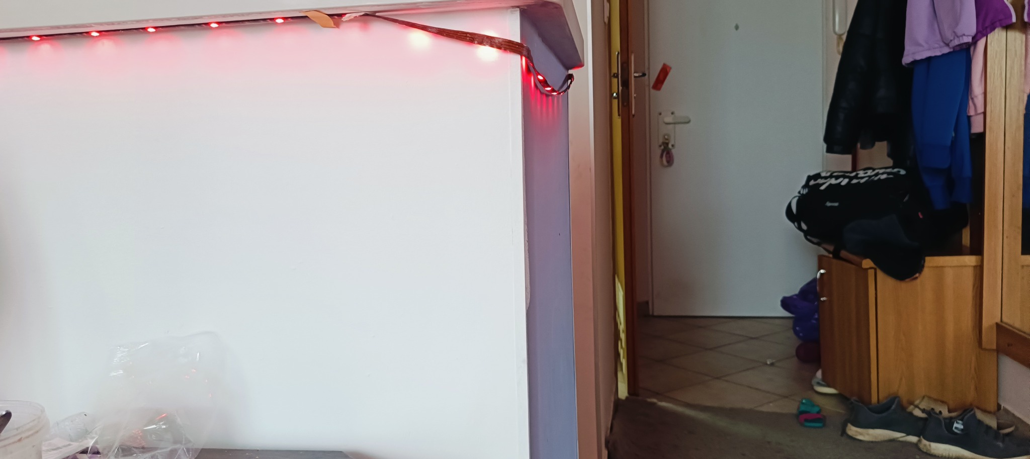 Biała ściana z czerwoną listwą LED w rogu pokoju. Widoczny fragment przedpokoju z szafką na buty i ubrania. Ujęcie z bliska, oświetlenie mieszane.