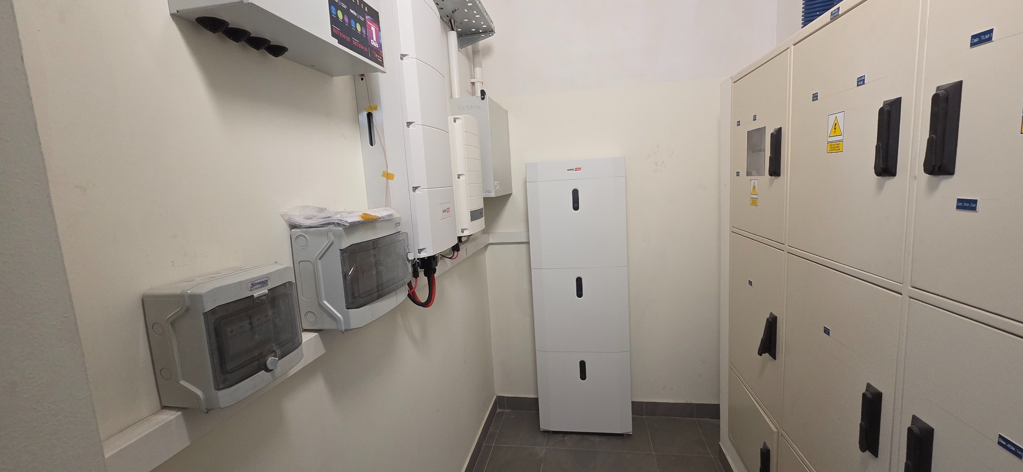Wnętrze pomieszczenia z zamontowanymi elementami instalacji fotowoltaicznej: rozdzielnice, inwerter i magazyn energii SolarEdge. Widoczne kable i oznaczenia ostrzegawcze.