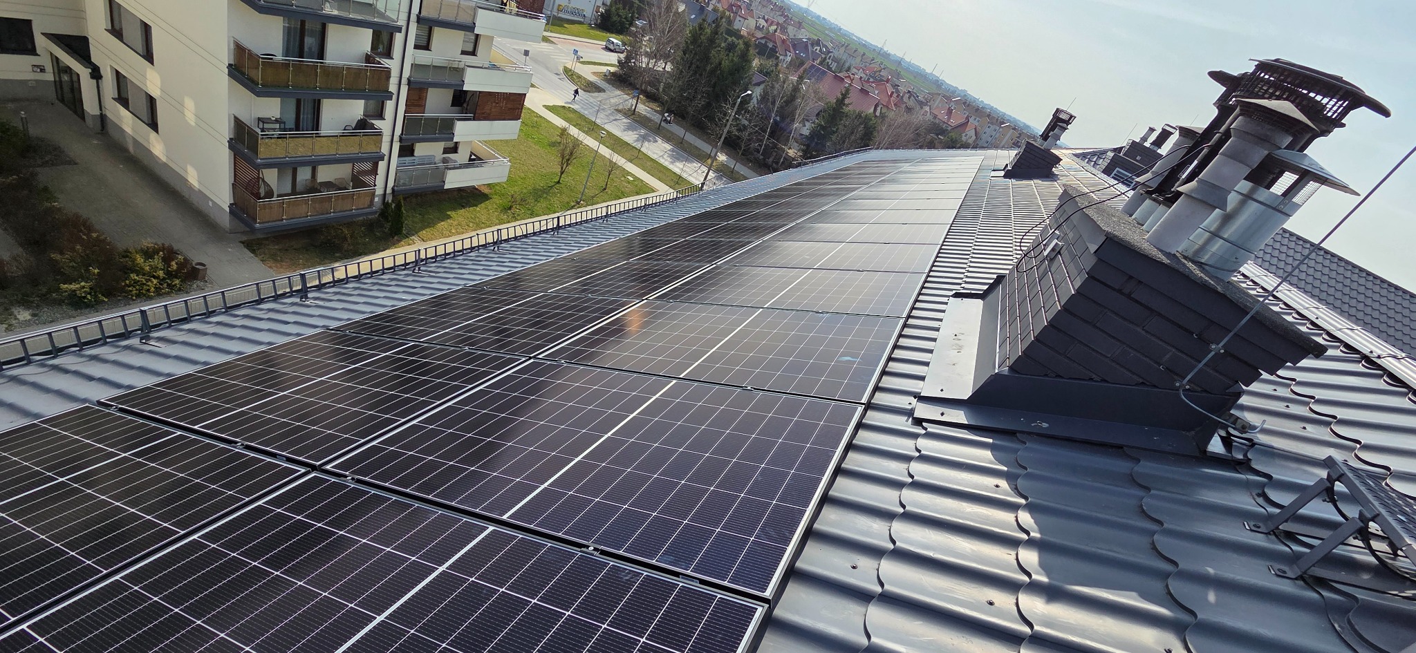 Panele słoneczne na dachu budynku wielorodzinnego, widoczne kominy i fragment osiedla w tle. Instalacja fotowoltaiczna na szarej dachówce.