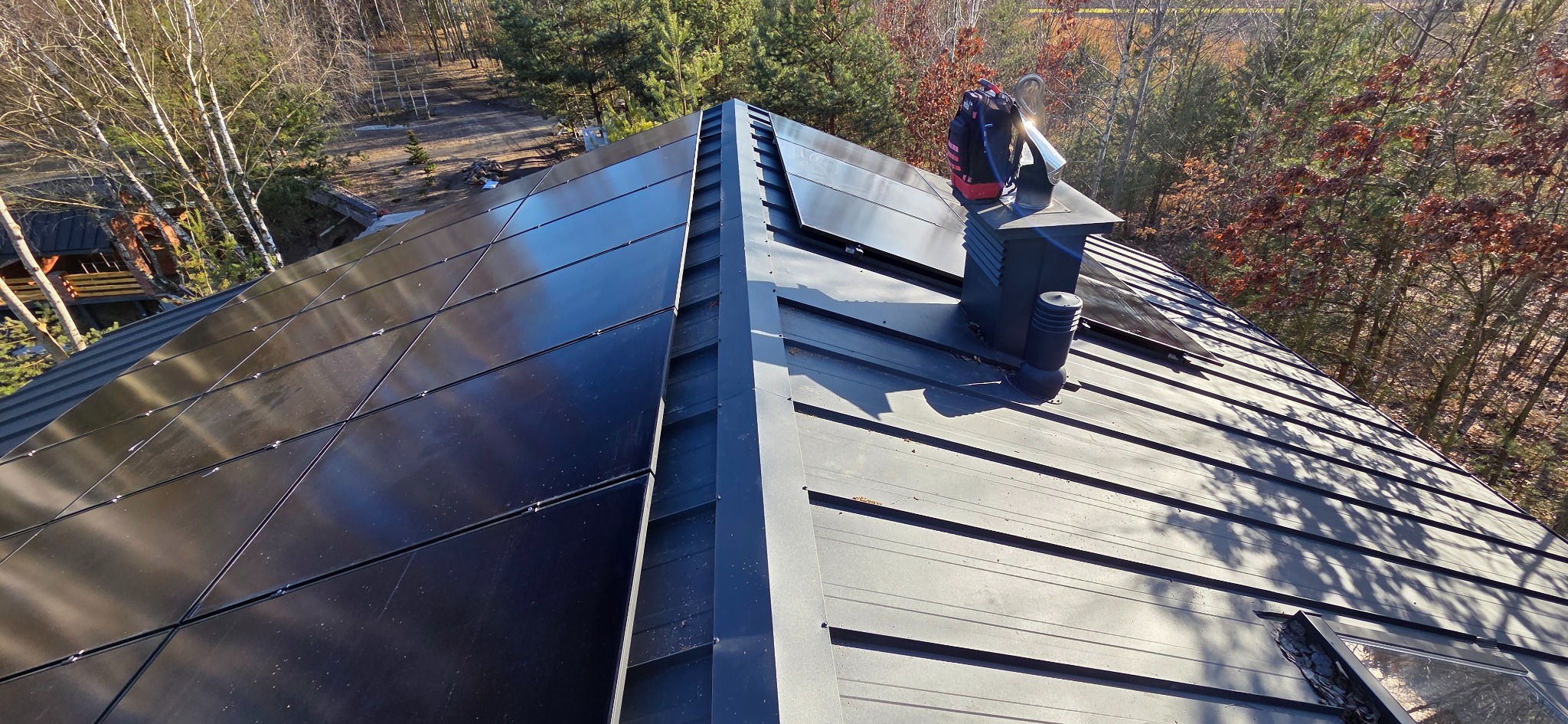 Czarny dach z panelami słonecznymi i kominem. Widok z góry, w tle drzewa. Część paneli jest zamontowana, a część jeszcze nie. Na kominie leży plecak.