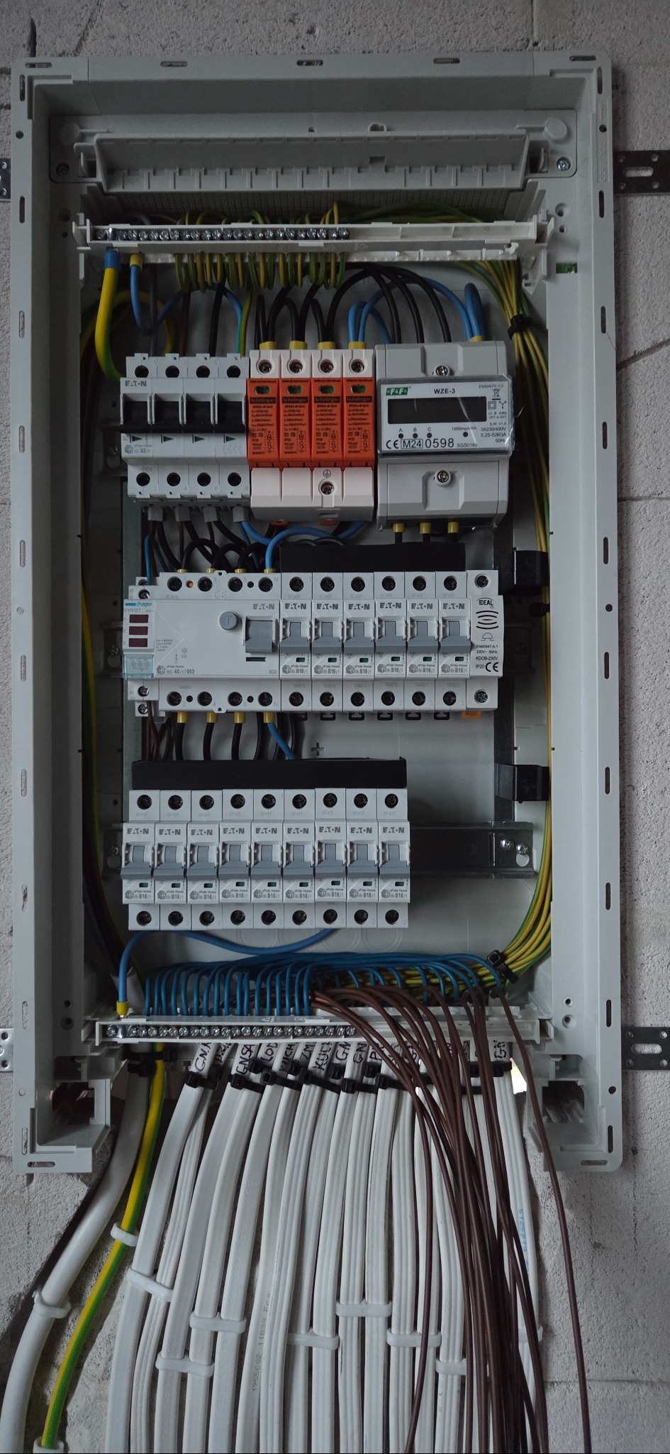 Rozdzielnica elektryczna z zabezpieczeniami przepięciowymi, bezpiecznikami i licznikiem energii. Widoczne kable oznaczone etykietami. Szara ściana w tle.