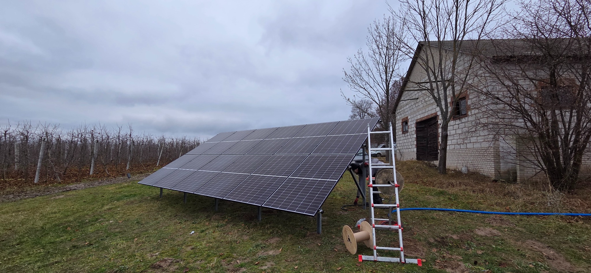 Panel fotowoltaiczny na trawie, drabina oparta o konstrukcję, budynek w tle. Instalacja w sadzie, pochmurne niebo. Widoczny bęben z kablem.