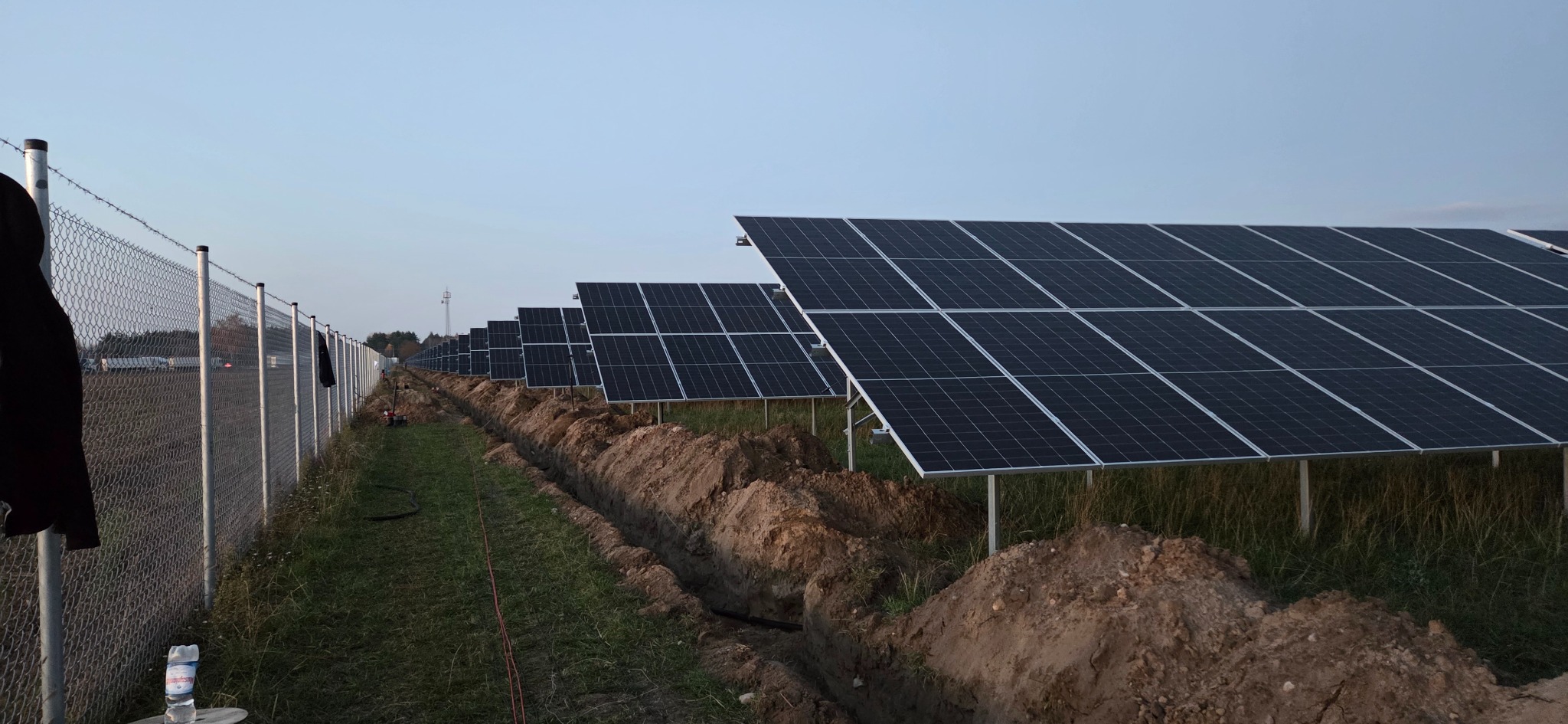 Rząd paneli fotowoltaicznych w trakcie instalacji, widoczne wykopy pod kable, ogrodzenie z siatki i butelka wody. Ujęcie z niskiej perspektywy.