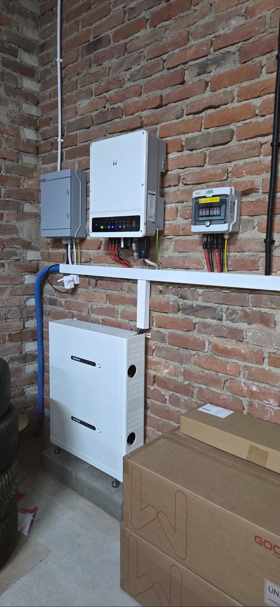 Instalacja magazynu energii GoodWe na ceglanej ścianie. Widoczne okablowanie, skrzynki i inwerter. Elementy systemu zamontowane na białej listwie. Część opon i kartonów w tle.