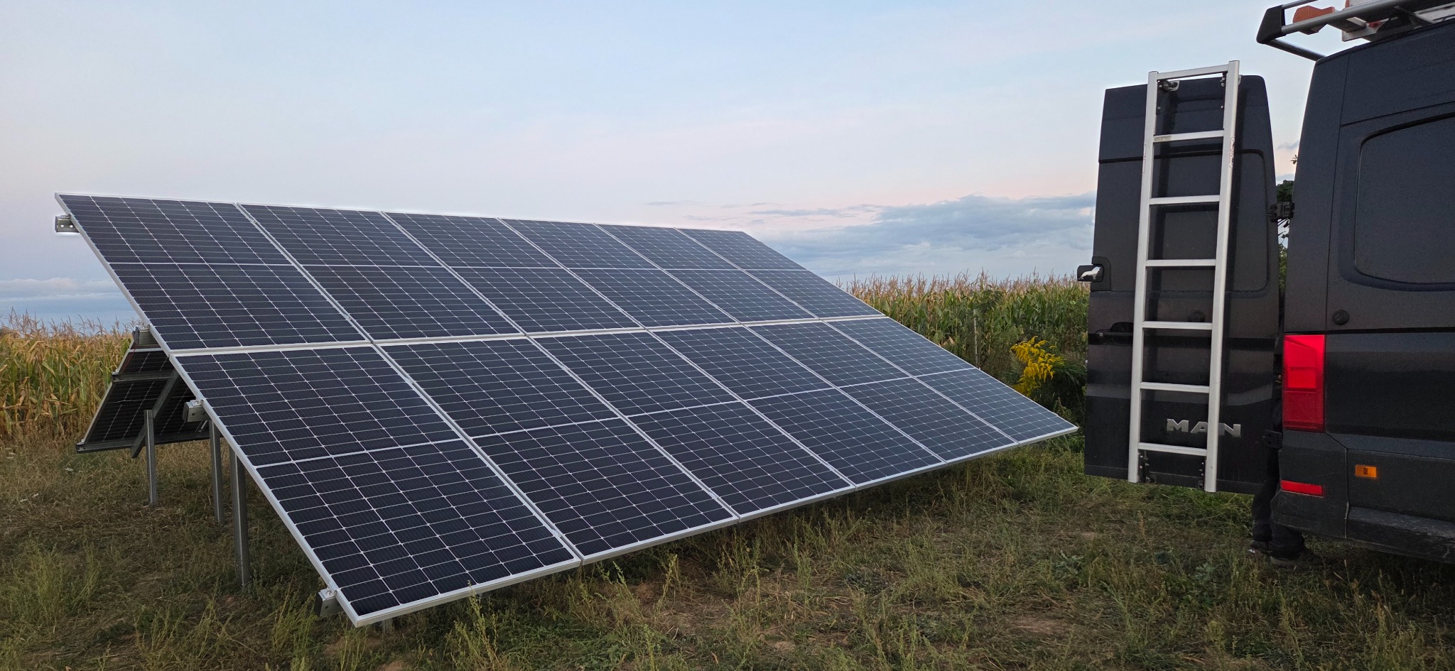 Panel fotowoltaiczny na łące, obok czarny bus z drabiną. Instalacja paneli na gruncie, w tle kukurydza i zachmurzone niebo. Nietypowe umiejscowienie paneli.