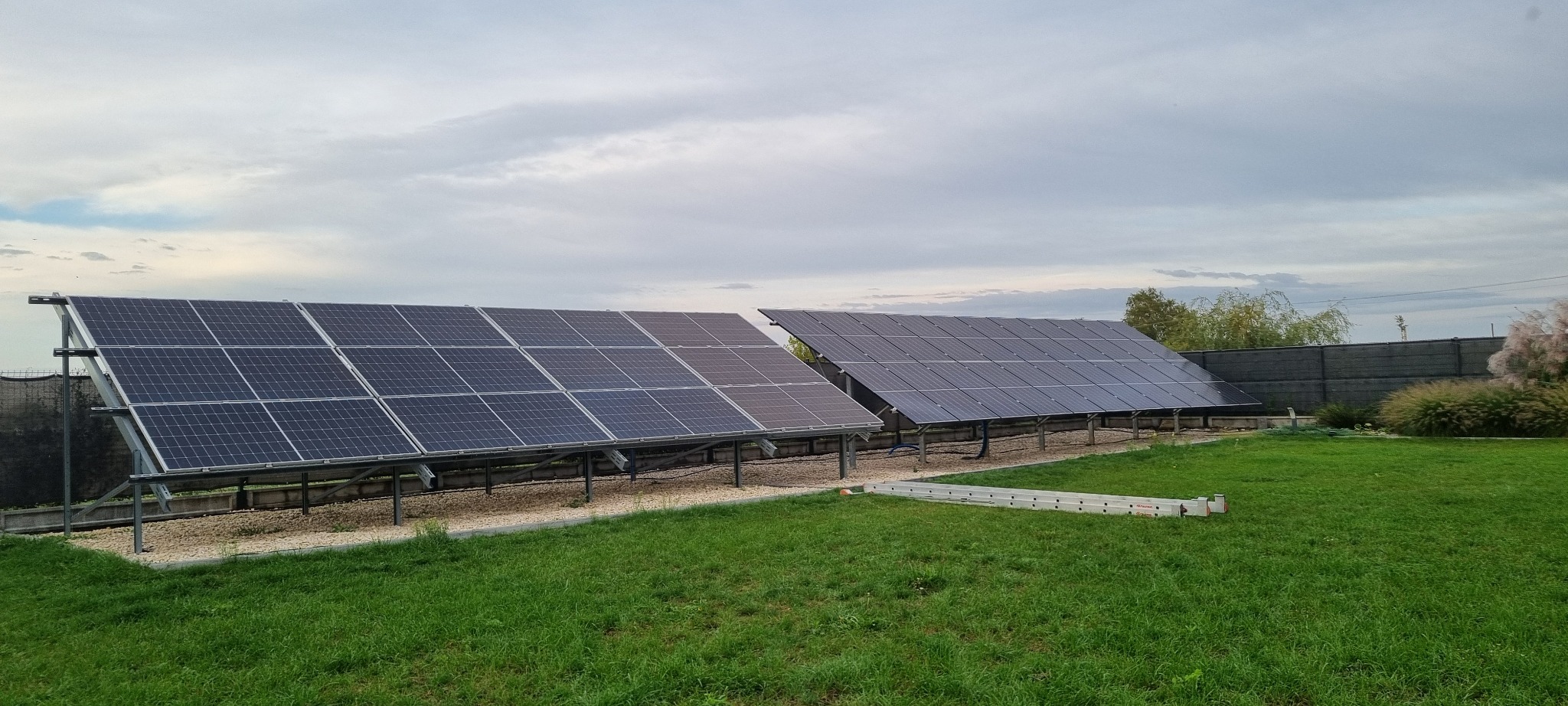 Dwa rzędy paneli słonecznych na metalowych konstrukcjach, ustawione na trawniku przed ogrodzeniem. Widoczne niebo z chmurami i drabina leżąca na trawie.