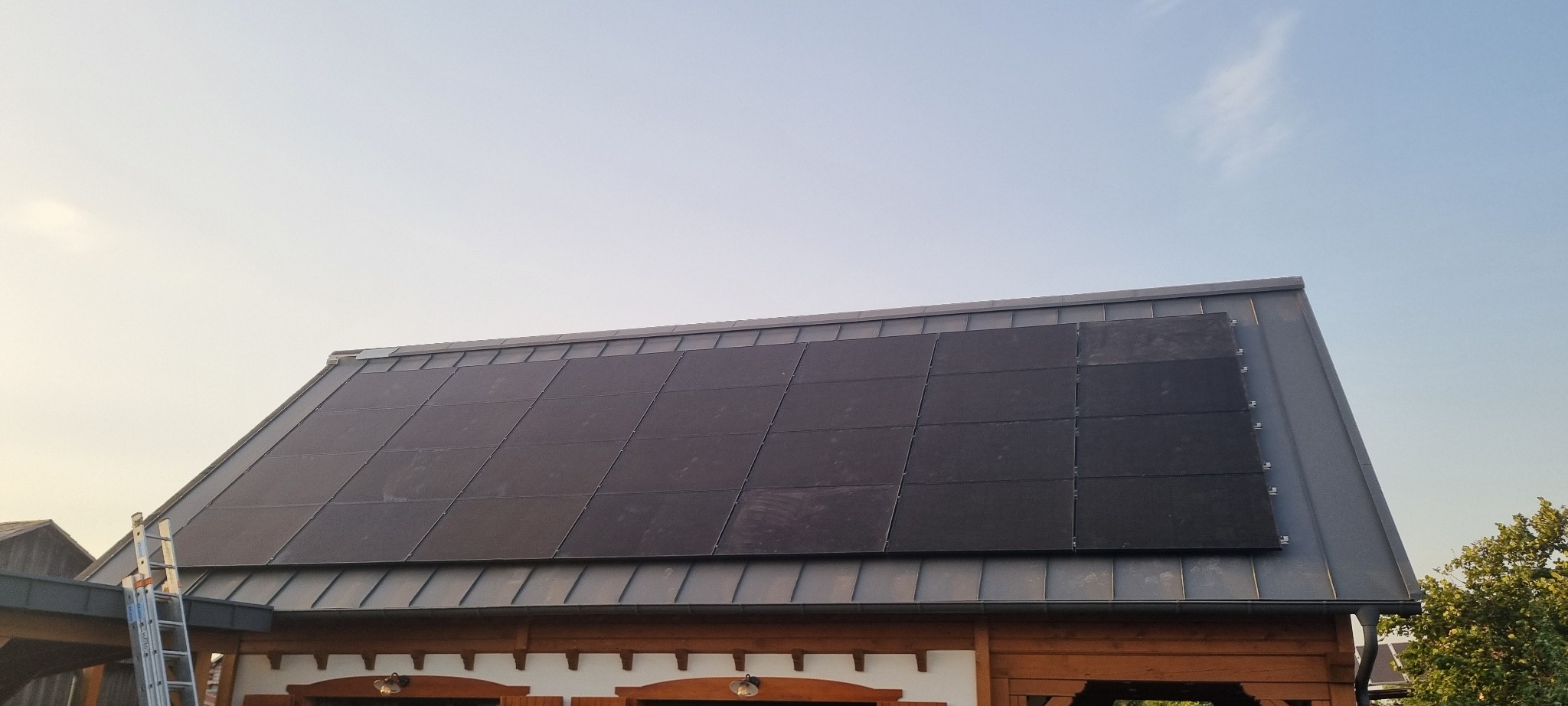 Panele słoneczne na dachu budynku z blachy, drabina oparta o rynnę, błękitne niebo w tle. Widok z dołu, lekko z lewej strony.