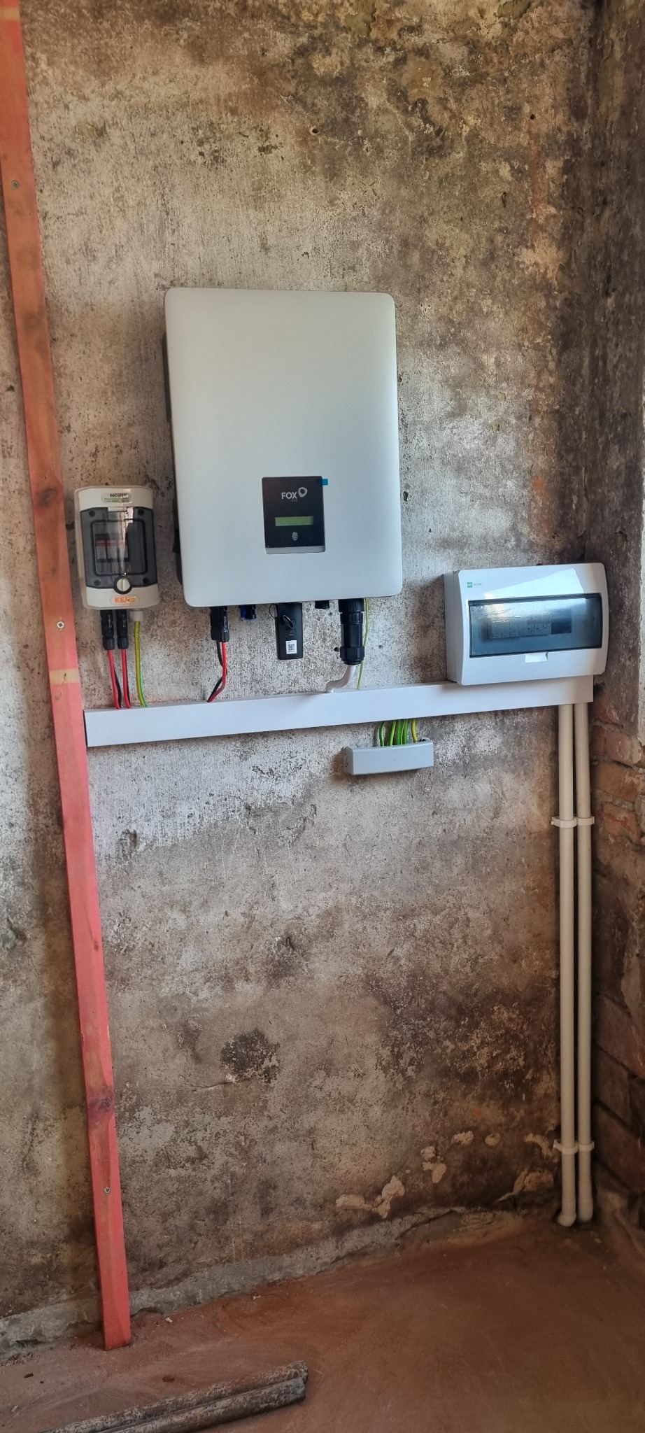 Zamontowany inwerter FoxESS, licznik energii i rozdzielnica na ścianie o surowym wykończeniu. Widoczne kable i rurki instalacyjne. Instalacja fotowoltaiczna wewnątrz budynku.