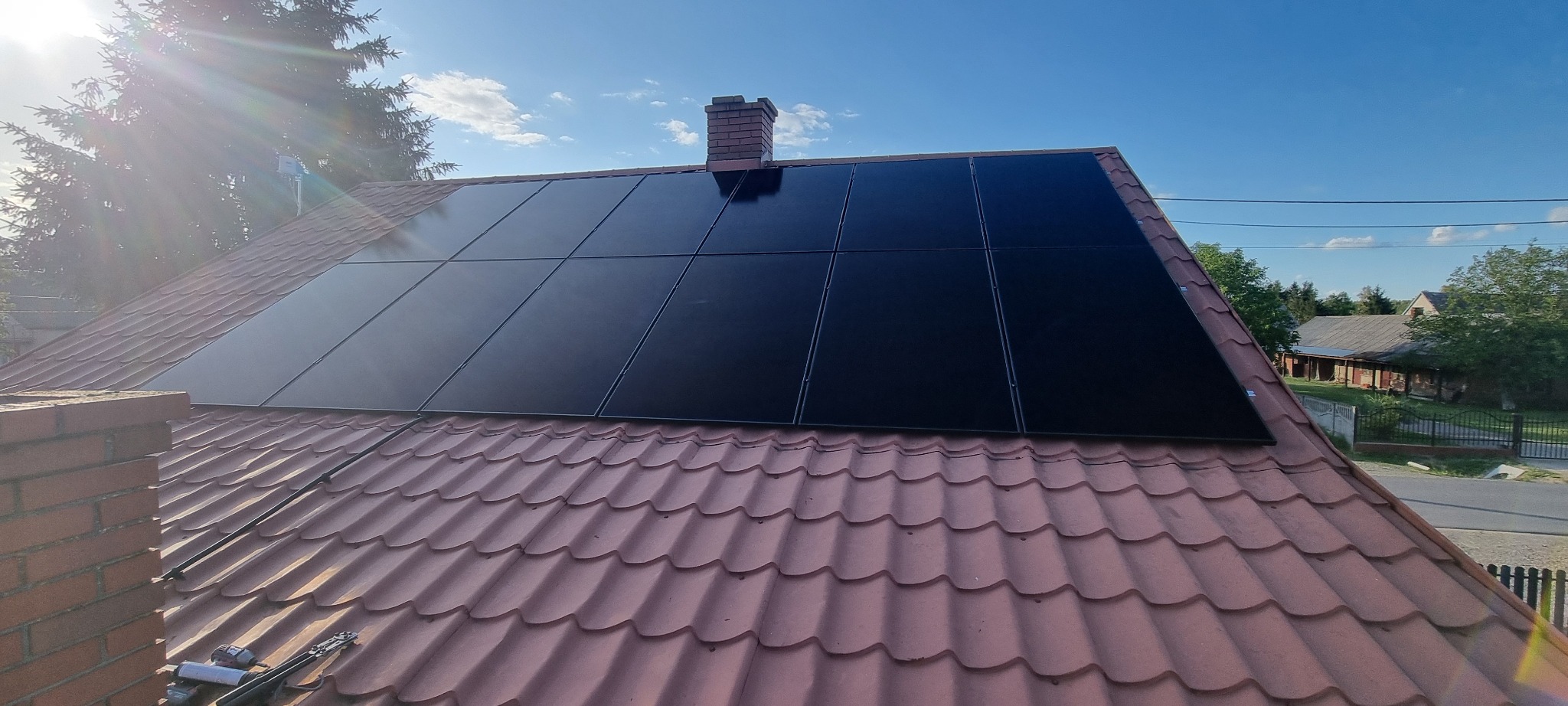 Błyszczące, czarne panele fotowoltaiczne na dachu z ceglastą dachówką, komin z cegły w tle, błękitne niebo i zieleń drzew w słoneczny dzień.