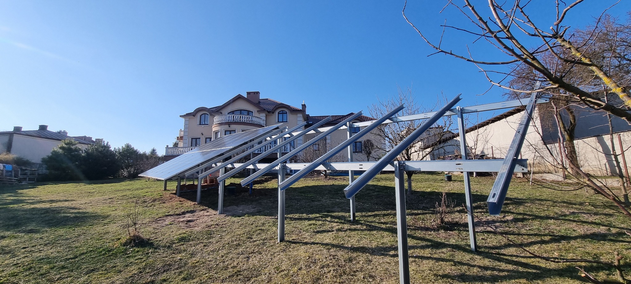 Montaż paneli słonecznych na metalowej konstrukcji na trawiastym terenie. W tle dom jednorodzinny i błękitne niebo. Częściowo zamontowane panele.